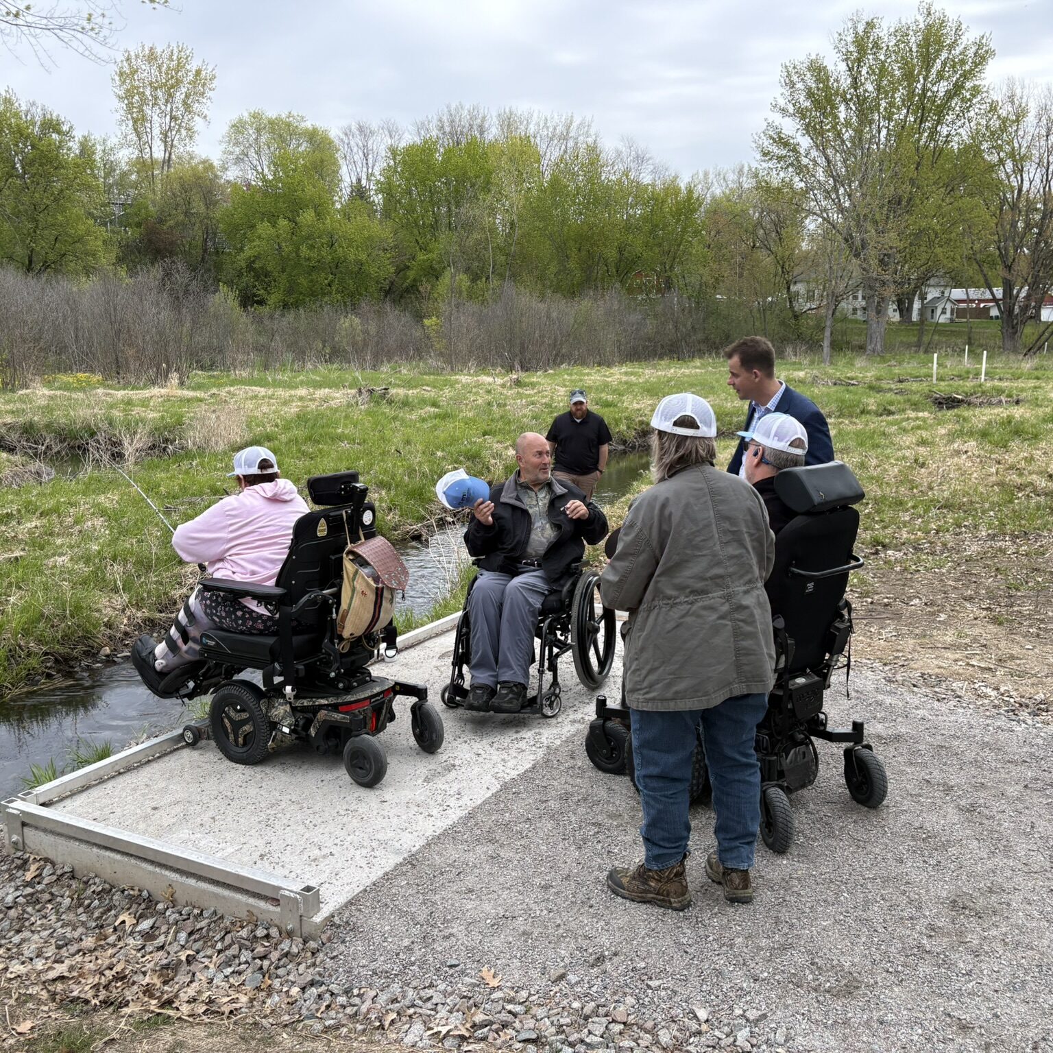 A group of people in a grassy area near a creek. There are three people in wheelchairs, one of whom is fishing.